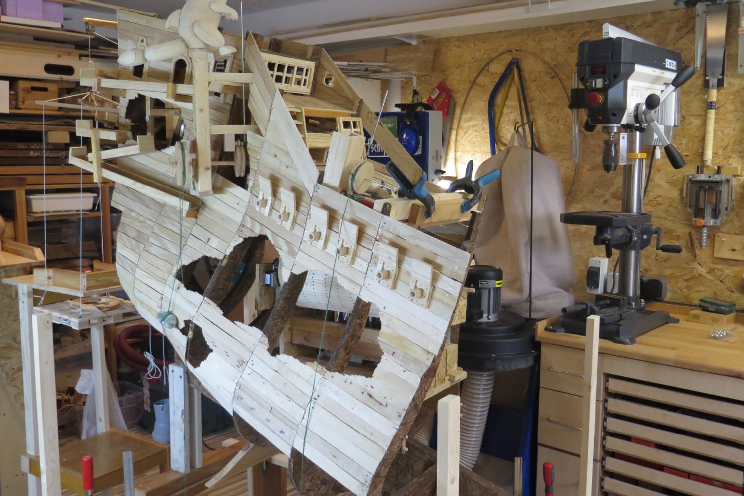 Ein großes, halbfertiges Holzmodell eines historischen Segelschiffs hängt stabil in einer Werkstattvorrichtung. Das Schiff zeigt viele handgearbeitete Details, etwa Fenster, Kanonenöffnungen und ein mehrstöckiges Heck. Die Umgebung ist eine Holzwerkstatt mit Werkzeugen, Maschinen und Holzmaterialien im Hintergrund.